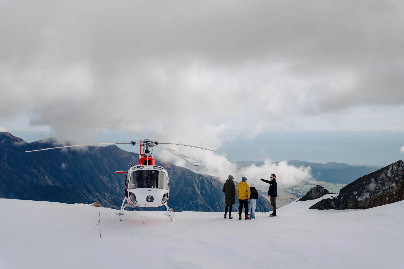 White helicopter on a snow ridge with four people watching clouds drift below