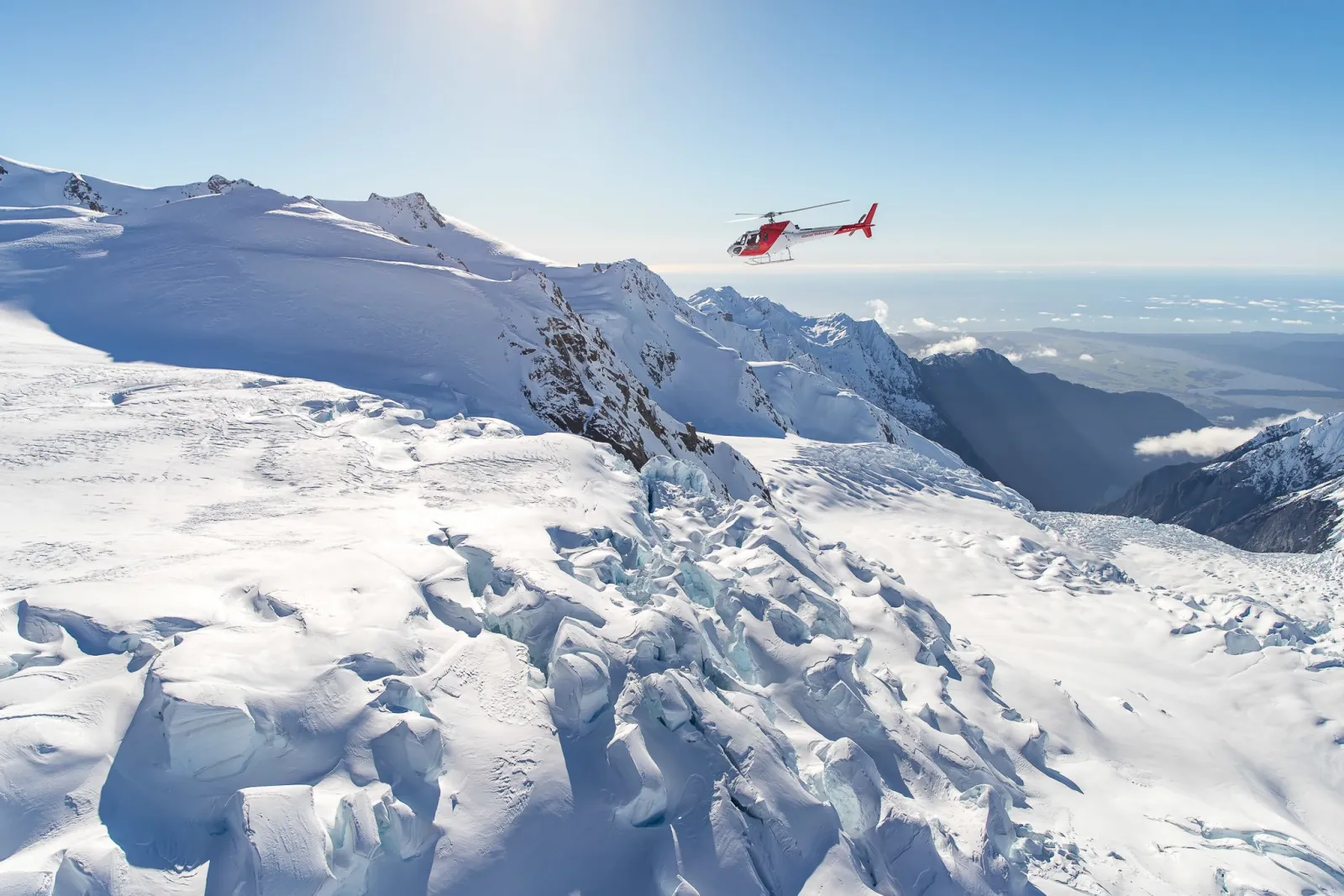 Small red helicopter flying over crevassed snow slopes with a coastline in the distance