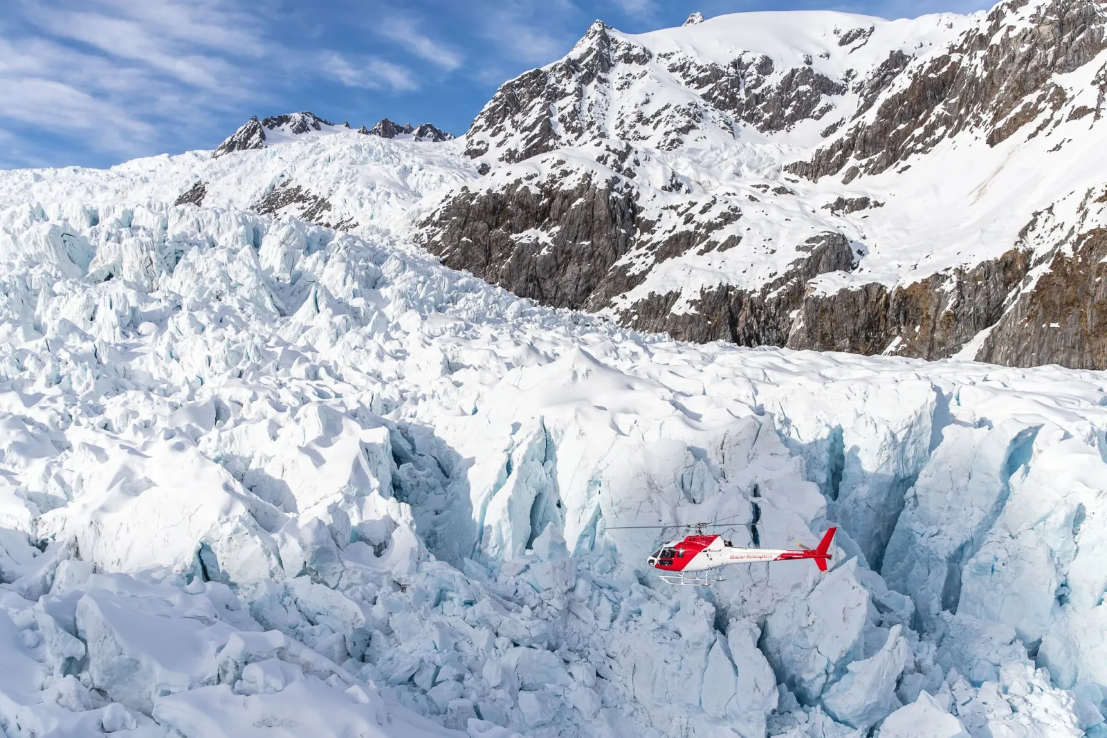 Small red helicopter beside a wall of blue-white glacier ice with a rocky peak above