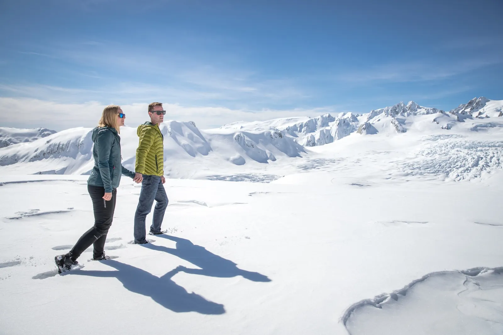 Man and woman walking across sunlit snow with a wide range of peaks behind them