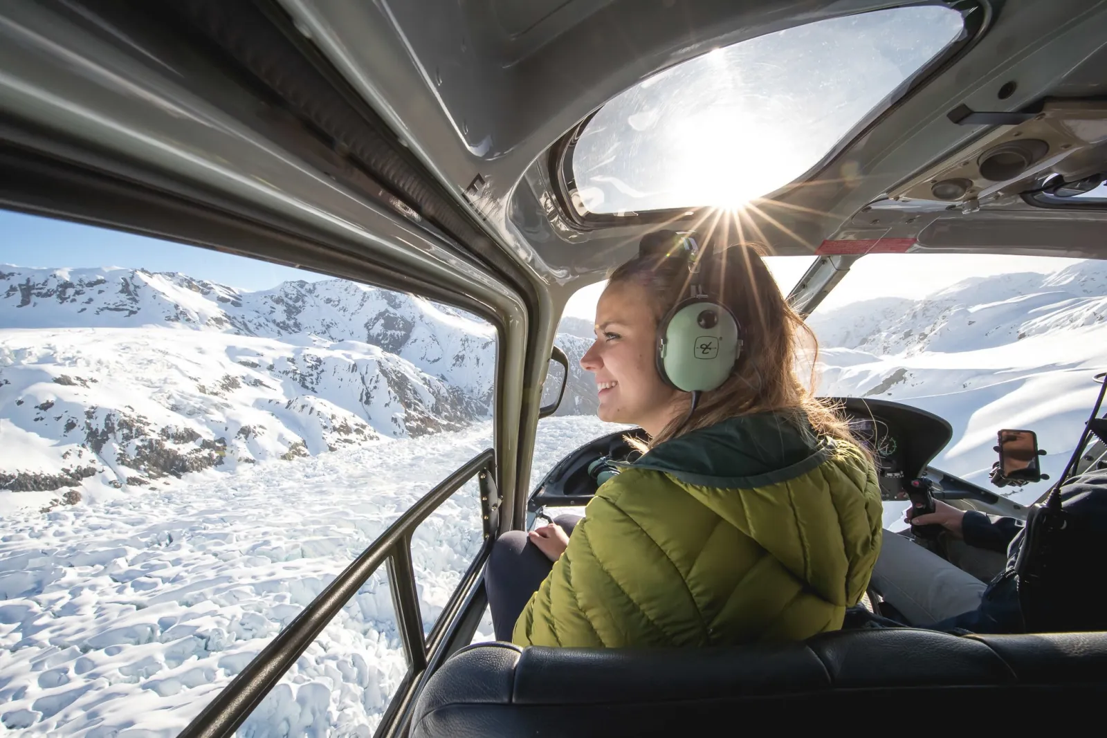 Woman in green jacket and headset smiling from an open helicopter door above snowy mountains