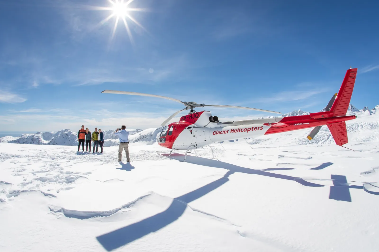 Red and white helicopter on a glacier with four people standing nearby in strong sun