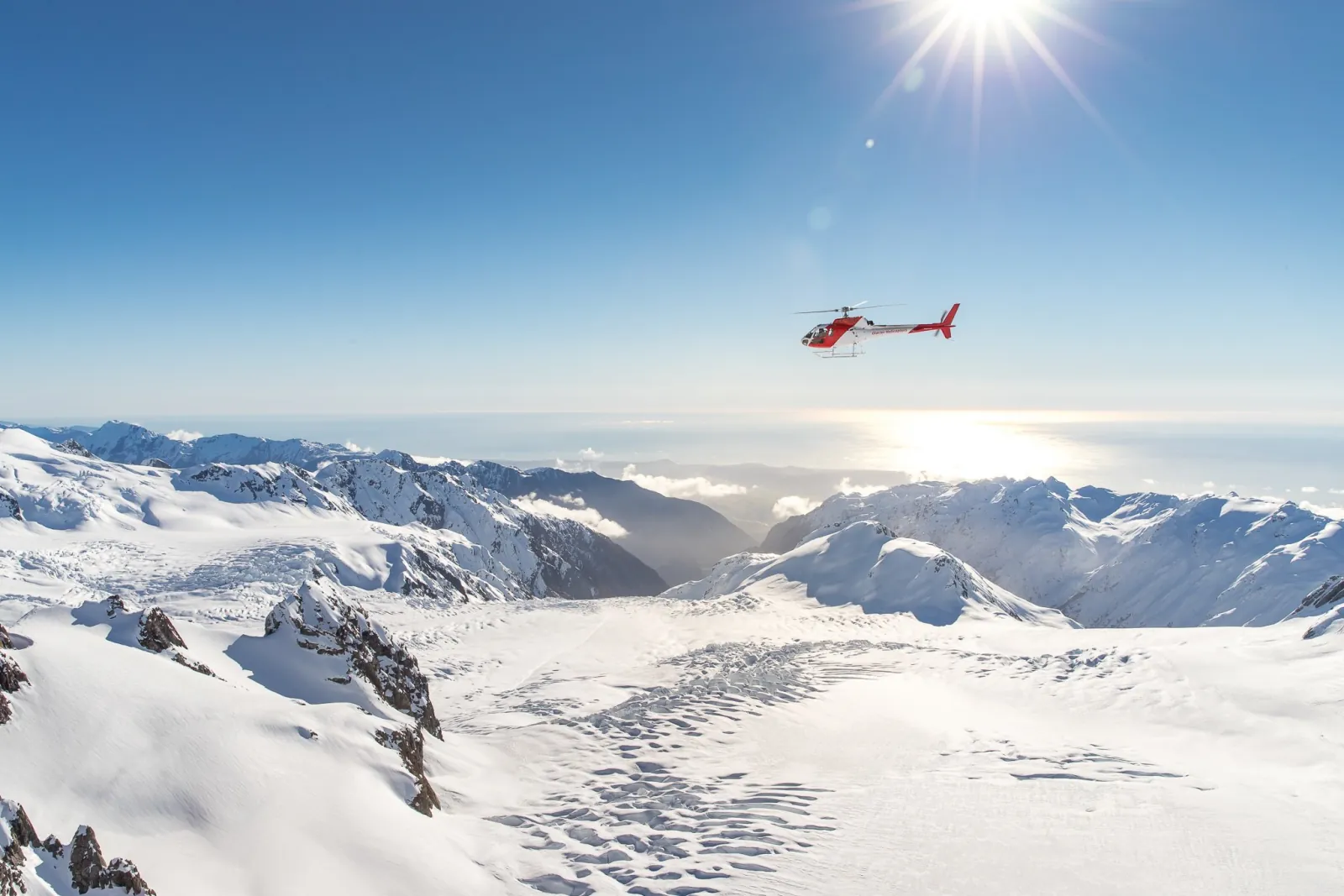 Red helicopter in flight over a wide snow basin with a bright sun and distant coastline