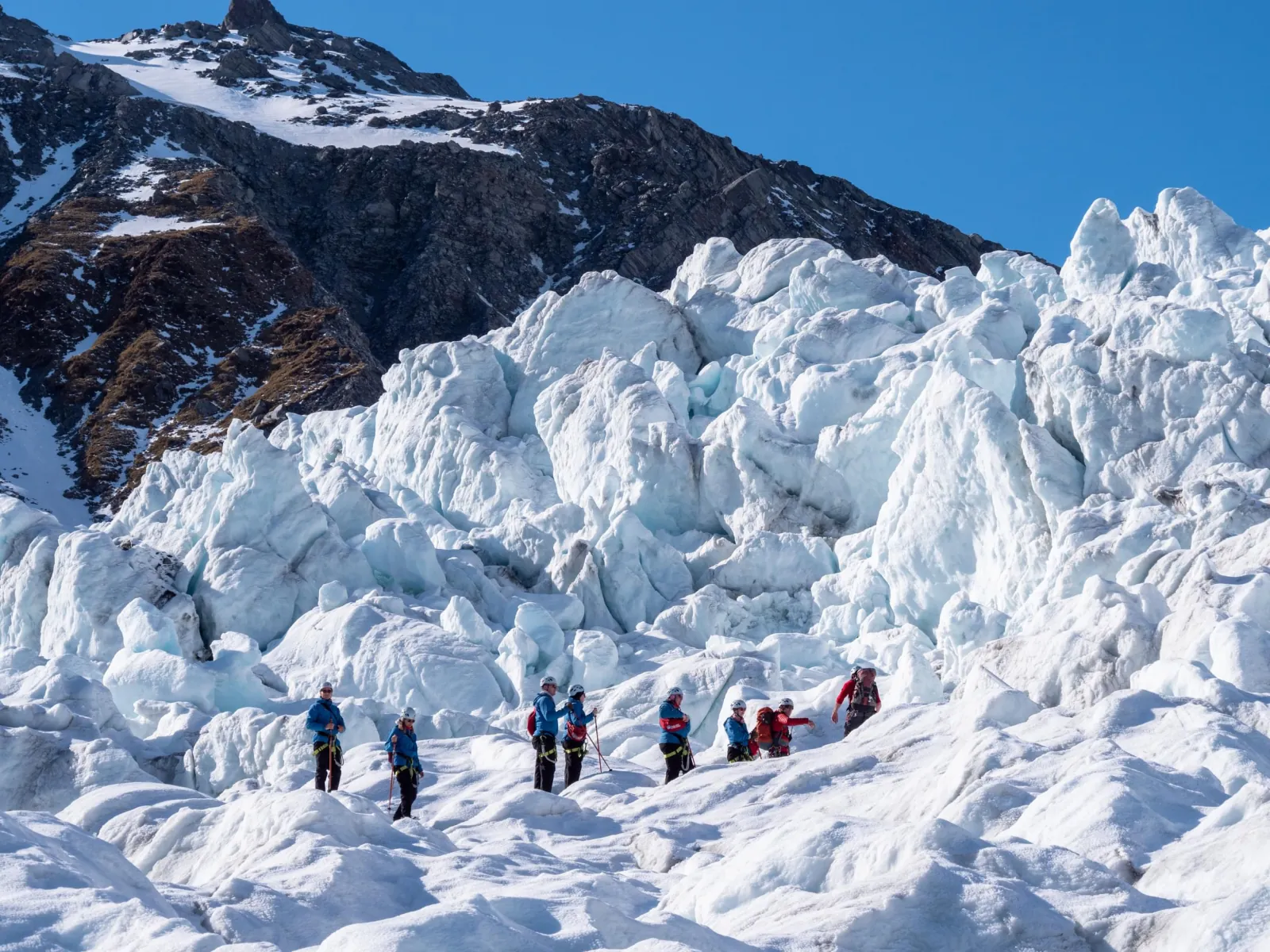 Line of tiny hikers crossing a rugged icefall beneath a dark rock face