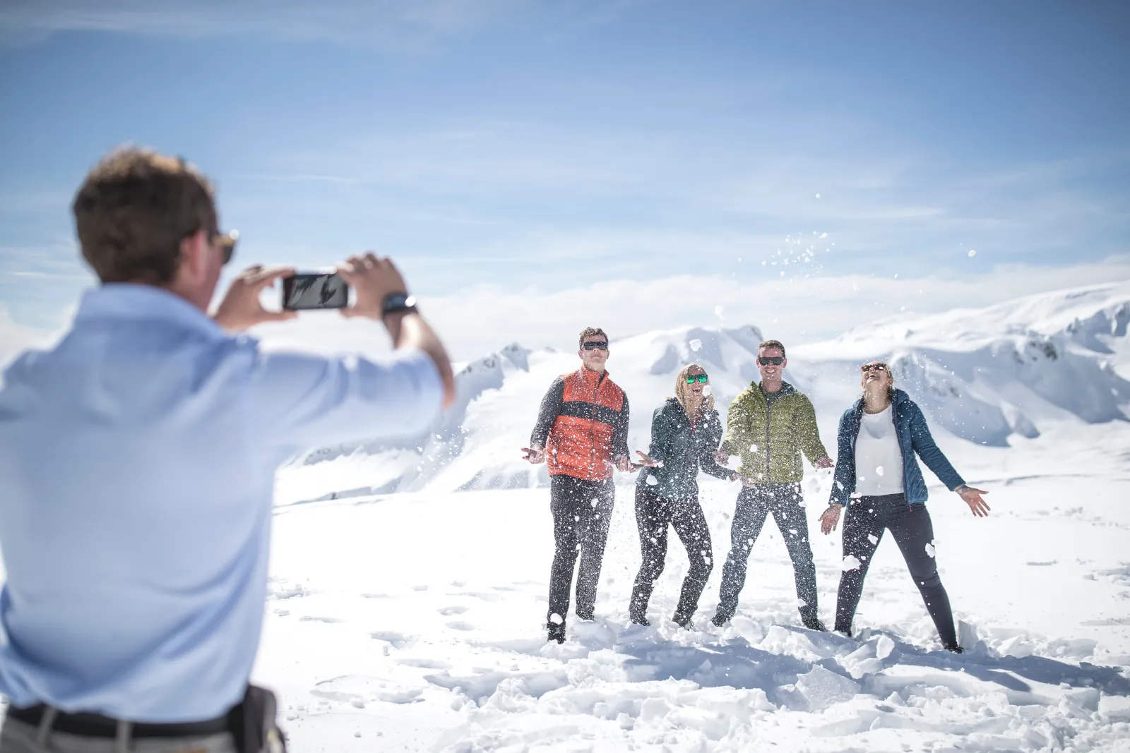 Four people laughing in a snow flurry while a man photographs them with a phone