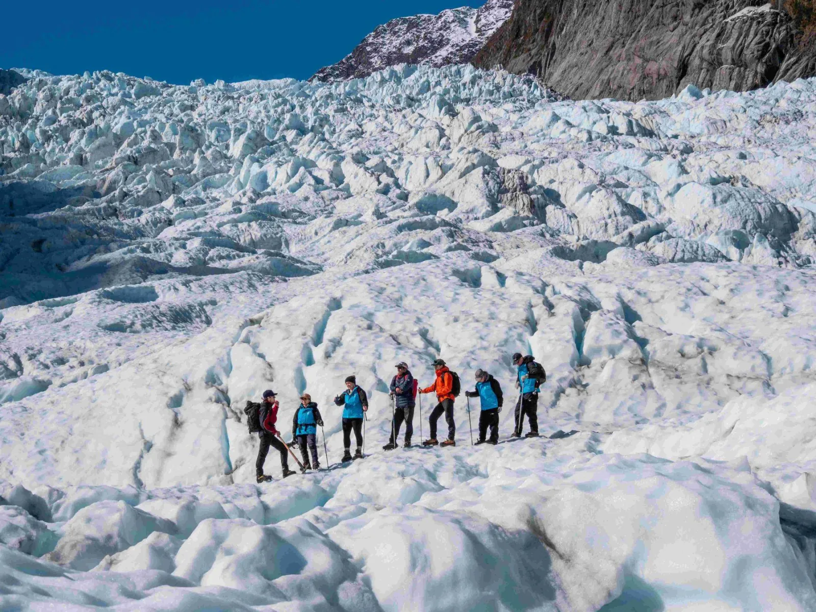 Long line of hikers with poles crossing a broken, textured glacier surface