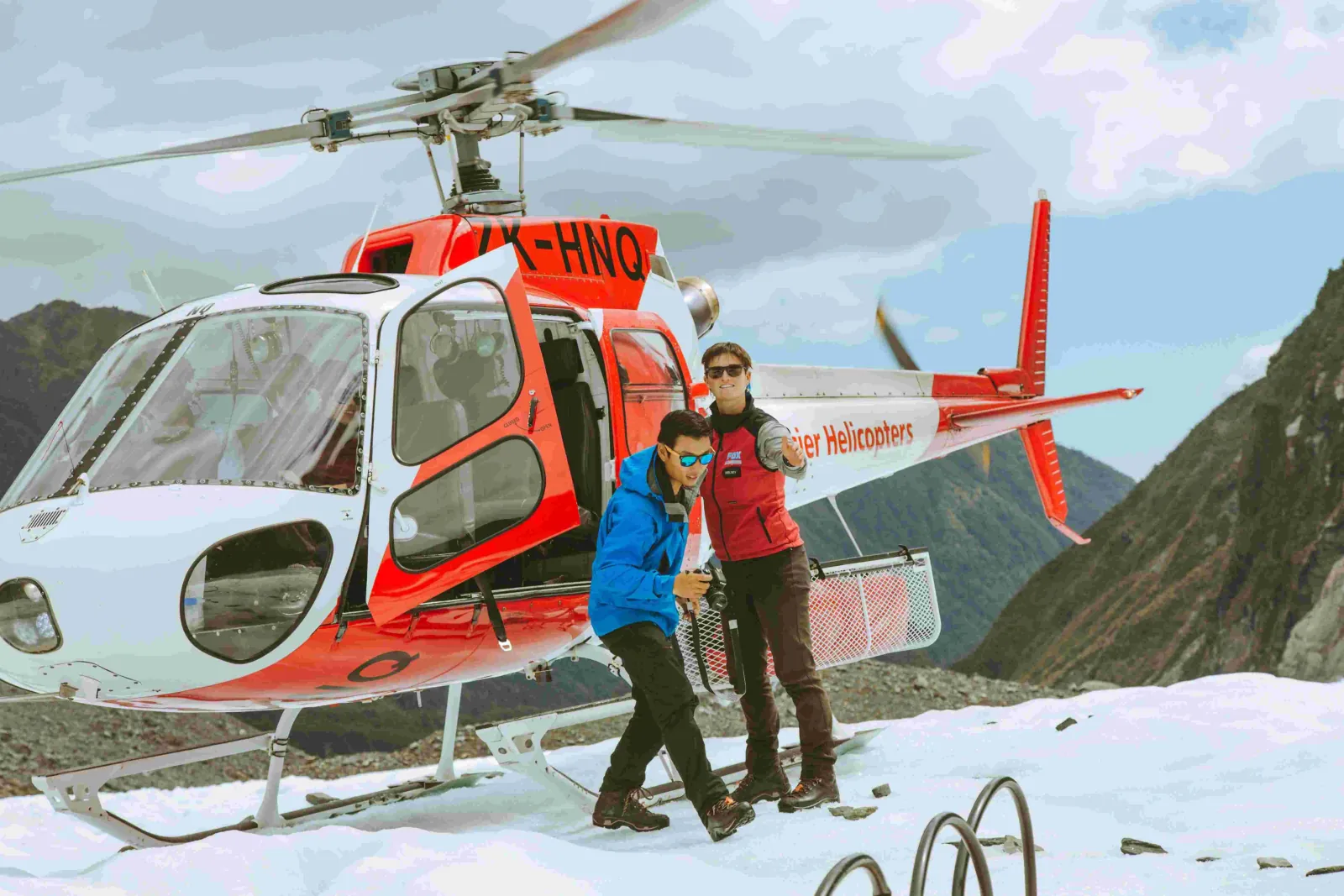 Pilot and passenger beside an open red and white helicopter on snow with green hills behind