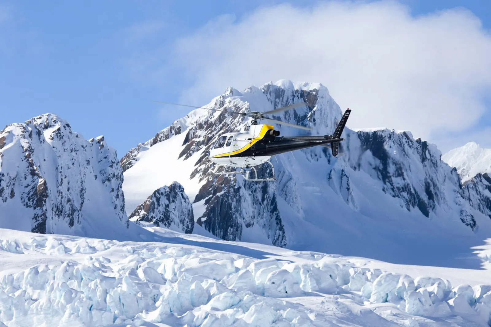 Yellow and black helicopter flying past a snow-covered rocky peak above crevassed ice