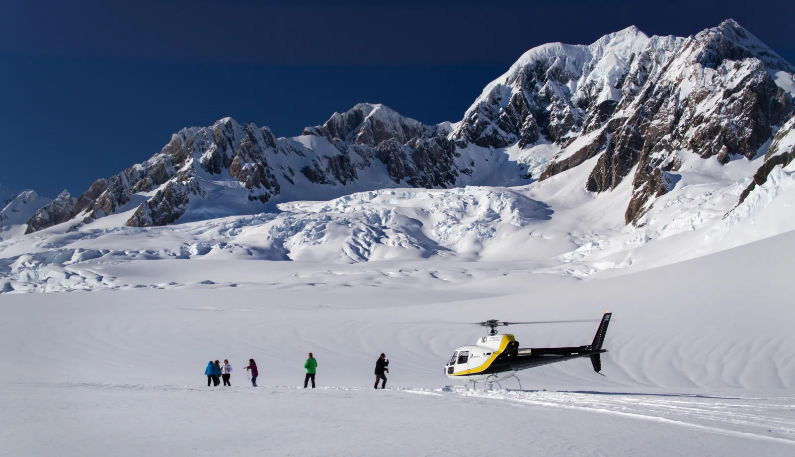 Yellow helicopter parked on a snowy plateau with tiny figures and jagged peaks above