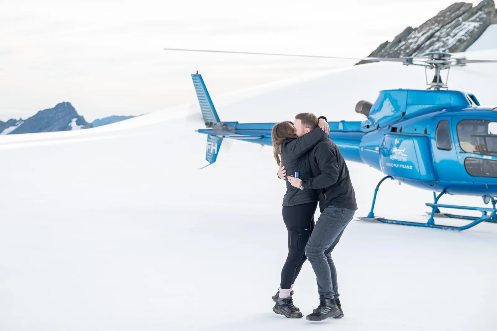 Couple embracing on a snowfield next to a blue helicopter with its rotor stopped
