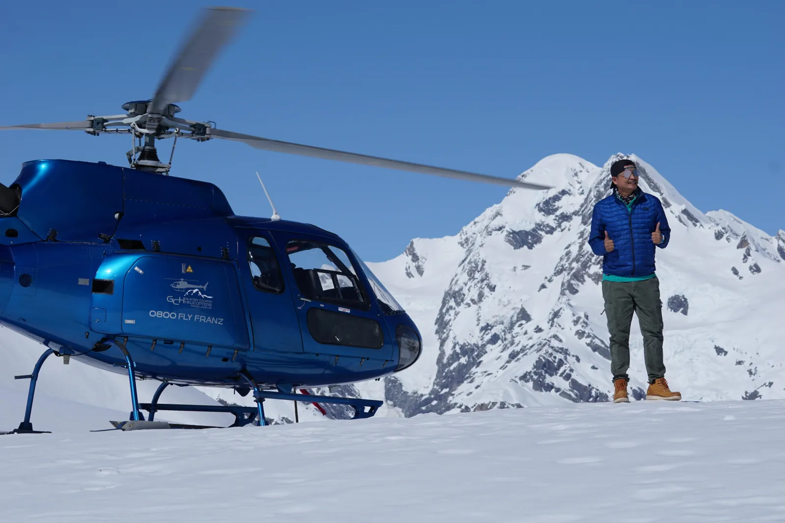 Man in blue jacket standing beside a blue helicopter on a snowfield below a peak