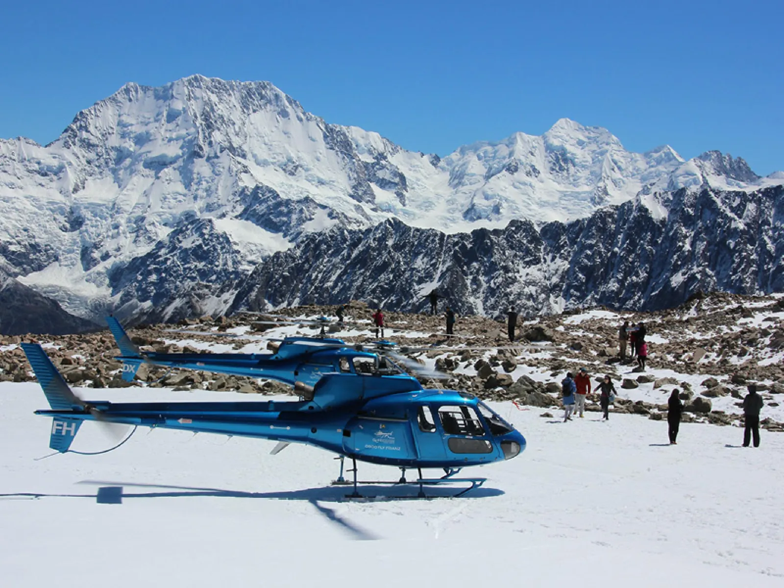 Blue helicopter on a snowfield with people on rocky ground below a large snowy peak