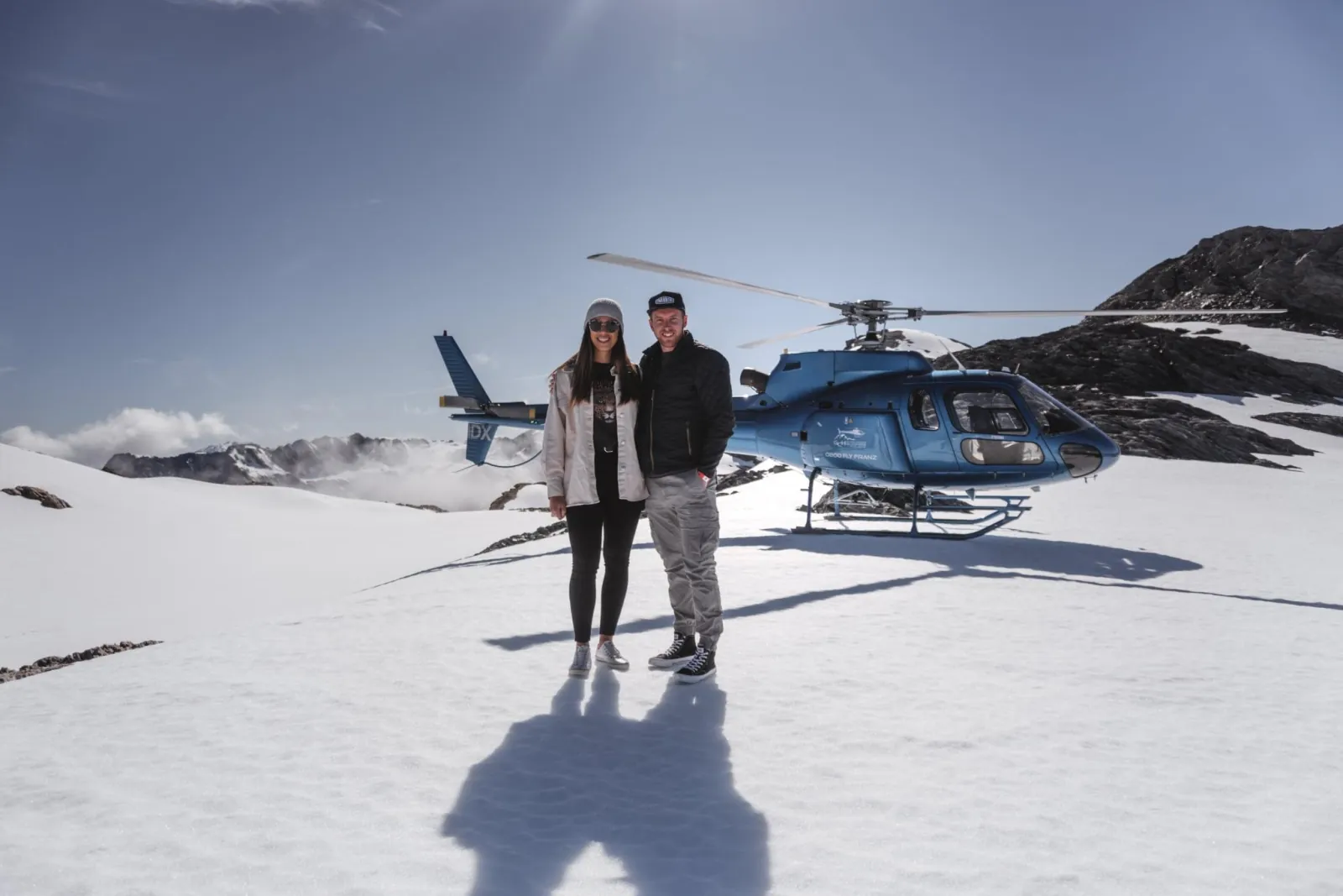 Couple posing on snow in front of a blue helicopter with long shadows