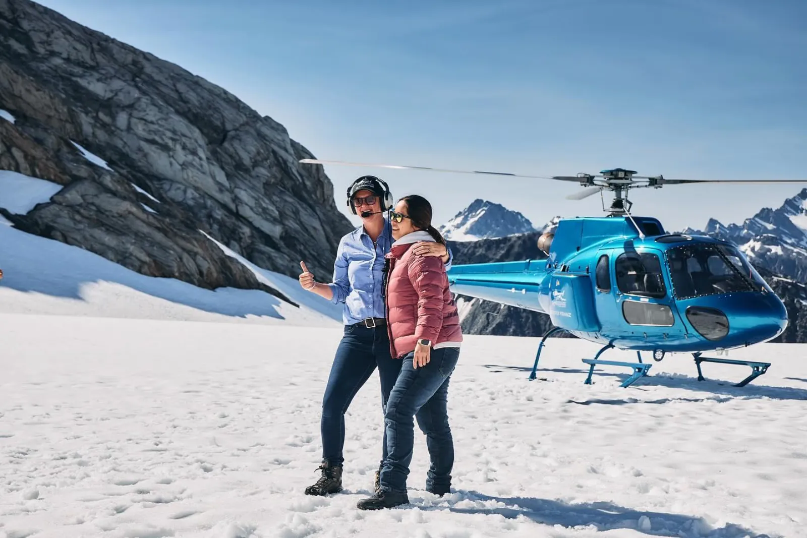 Two women posing in front of a turquoise helicopter with a rocky cliff behind