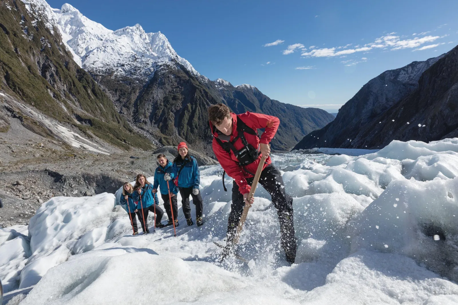 Guide in red jacket chipping ice while a group stands behind on glacier with mountains