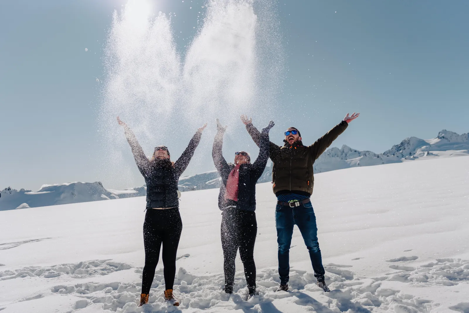 Three people jumping with arms raised as snow sprays upward on a glacier