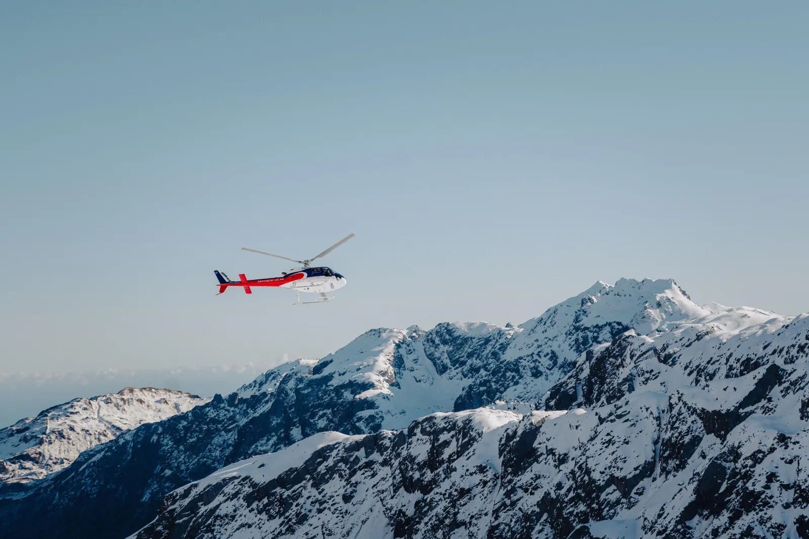 Red and blue helicopter flying above a snowy mountain ridge against pale sky