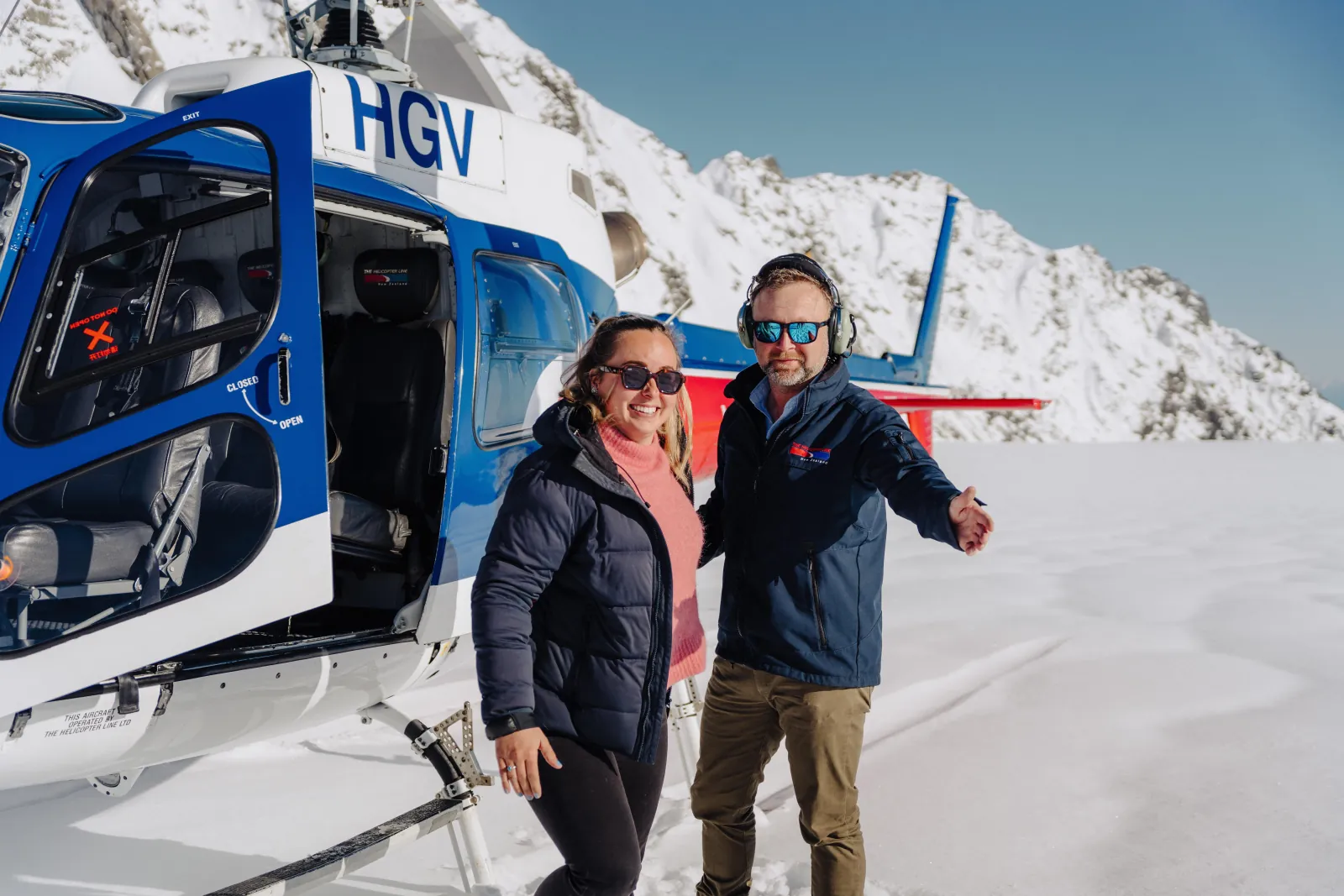 Smiling couple in jackets and headsets posing at the open door of a blue helicopter