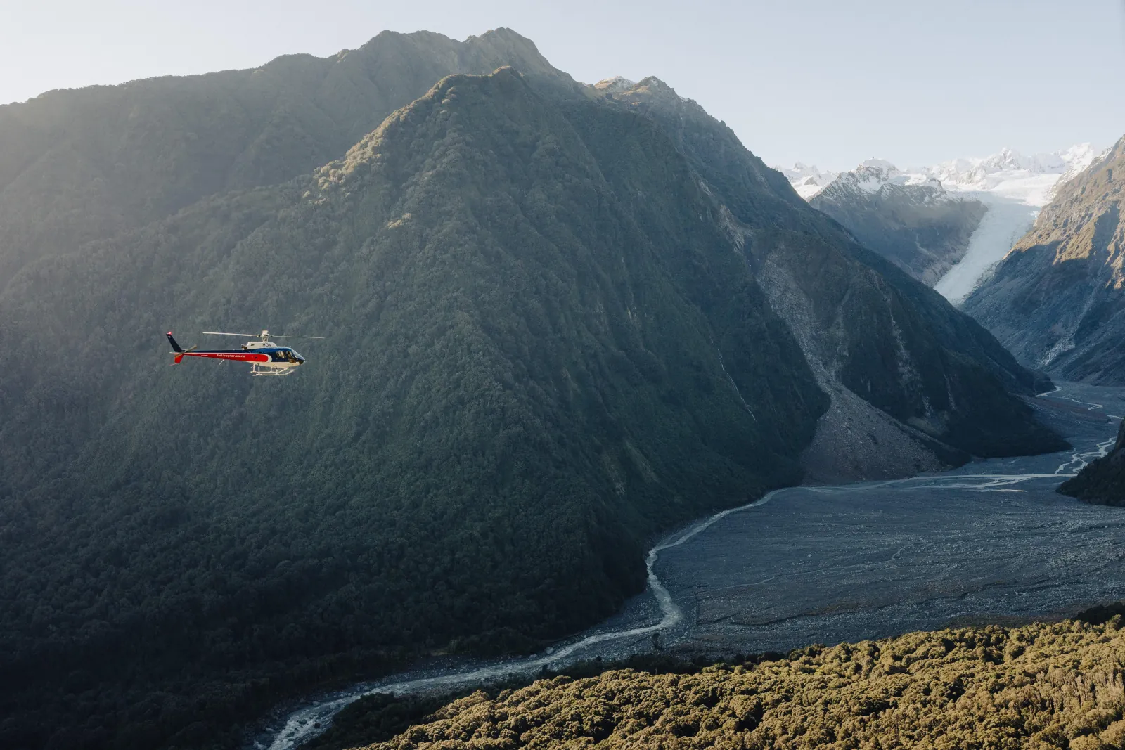 Red helicopter flying above a forested mountain valley with a braided river below