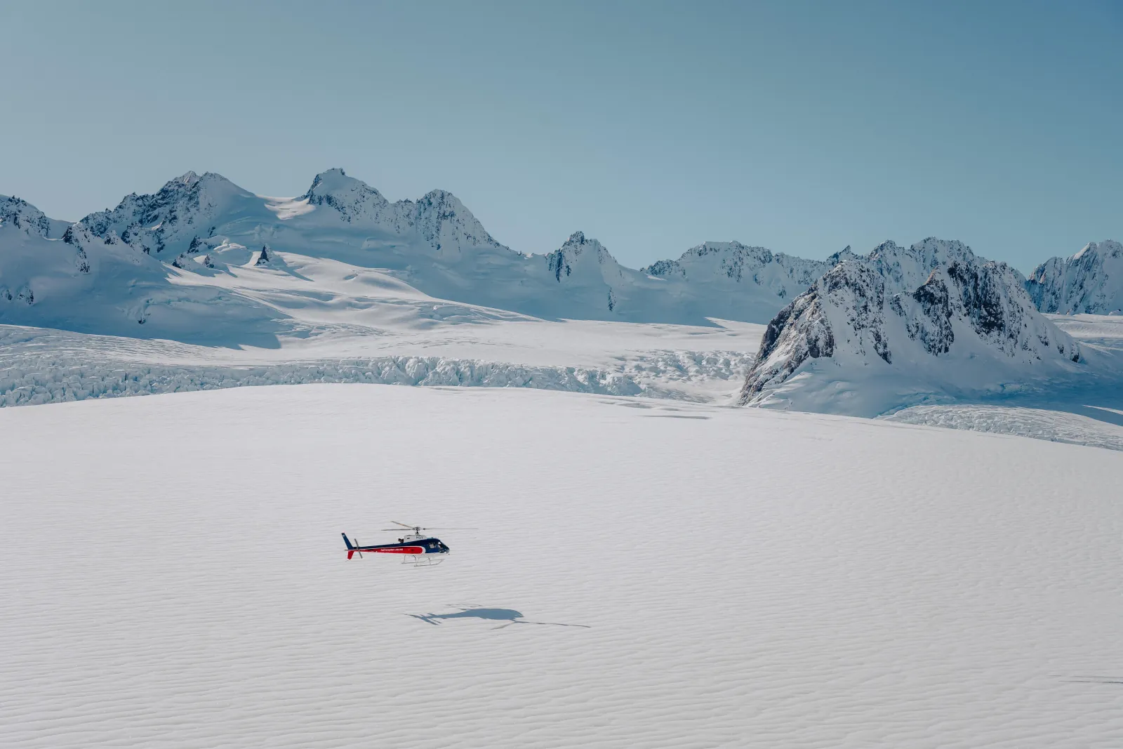 Tiny red helicopter and its shadow over a vast flat snowfield with peaks beyond
