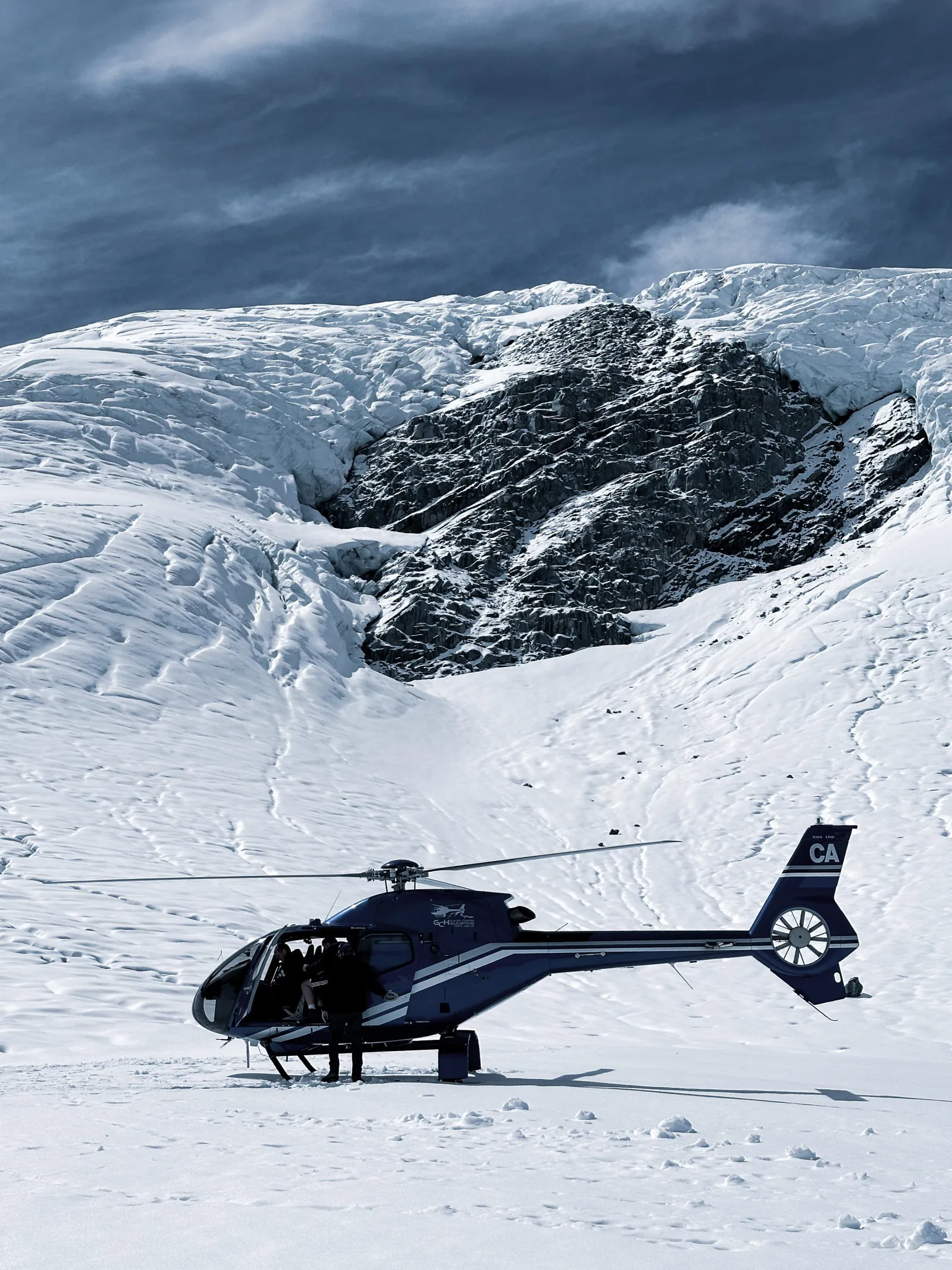 Dark helicopter landed on snow beside a rocky outcrop under heavy cloud