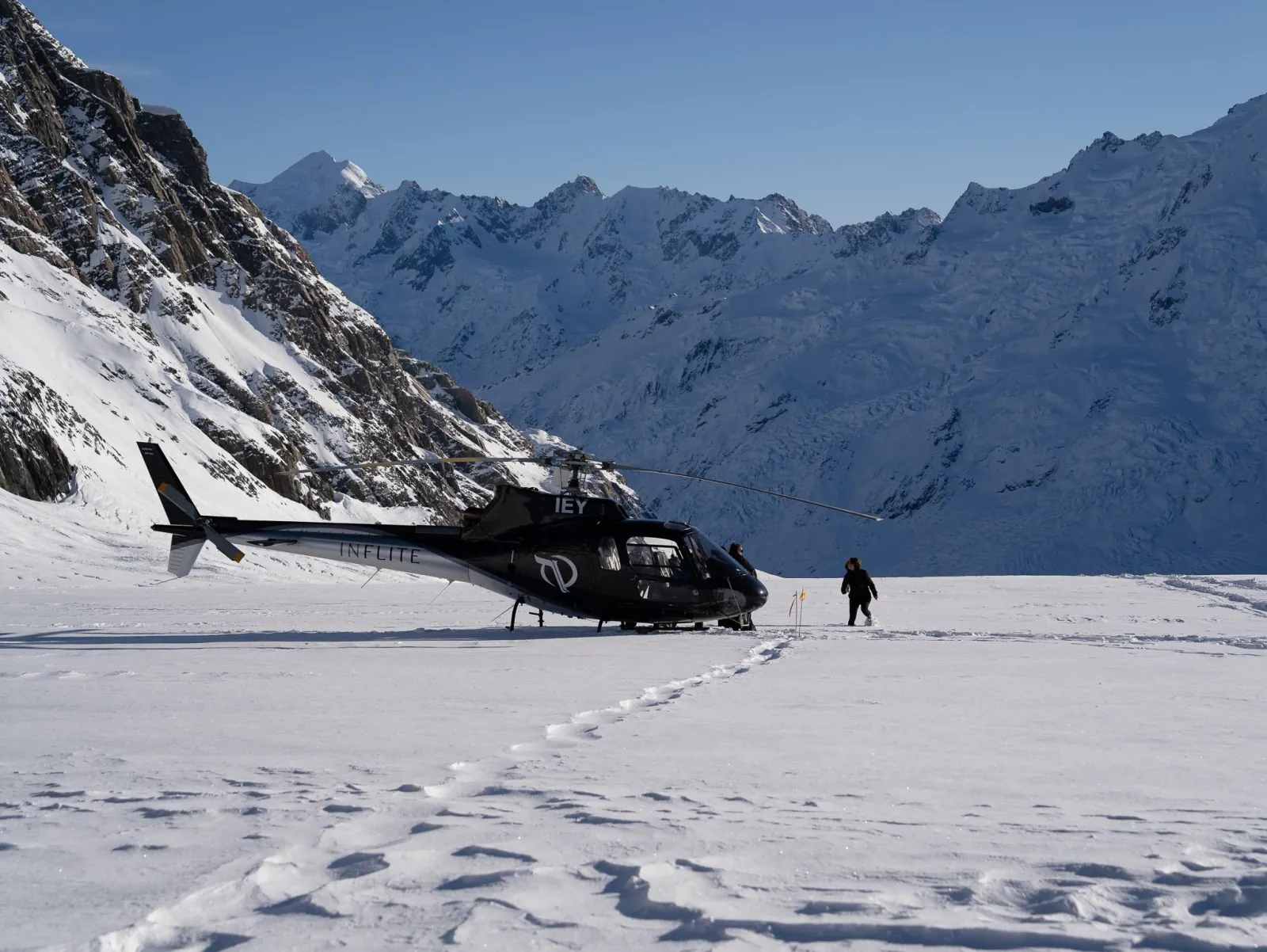 Black helicopter parked on a snowfield with one person walking toward valley peaks