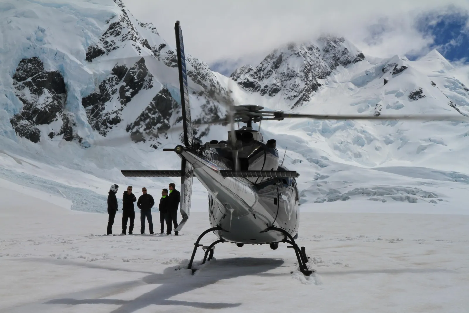 Grey helicopter on a glacier with four people standing in front, cloud on the peaks