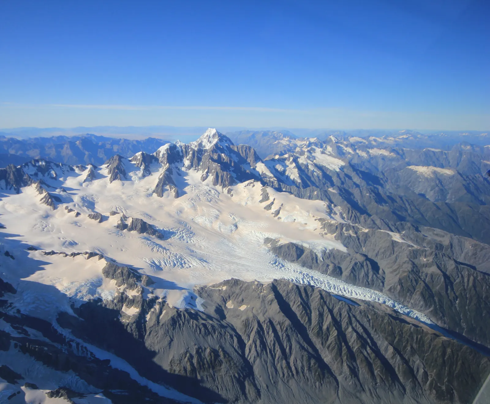 Aerial panorama of glaciers and ridges leading to a tall peak under clear blue sky