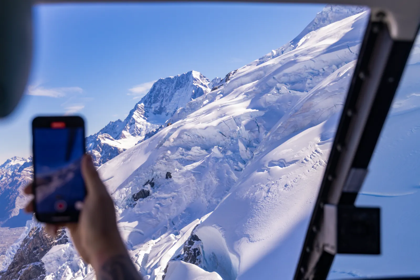 Hand holding a phone out of a helicopter window toward a big snowy mountain
