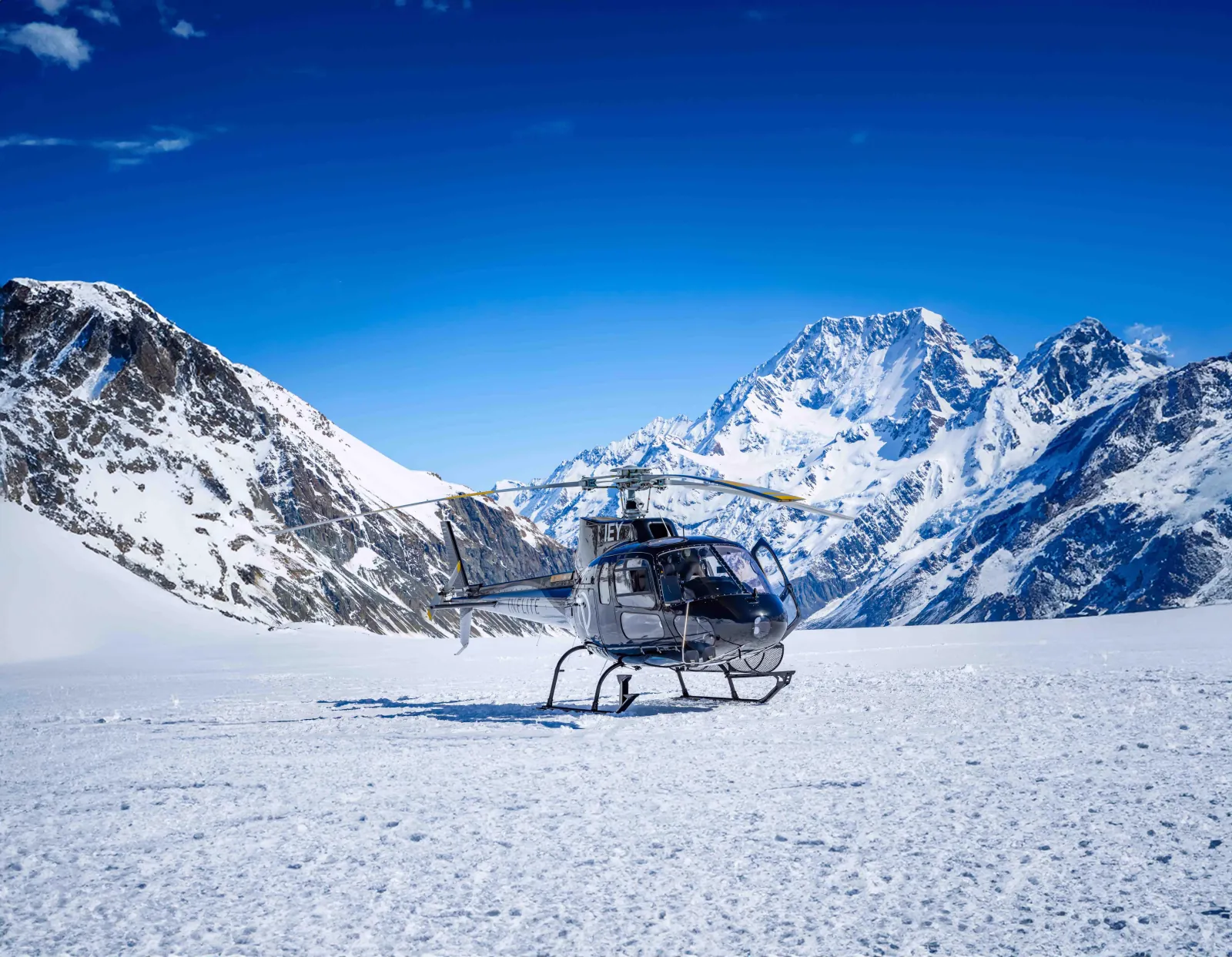 Grey helicopter landed on a snowfield with a high peak and ridges in the background