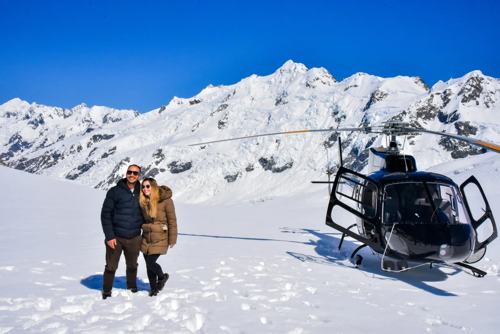 Couple standing close in snow beside a black helicopter with a mountain behind