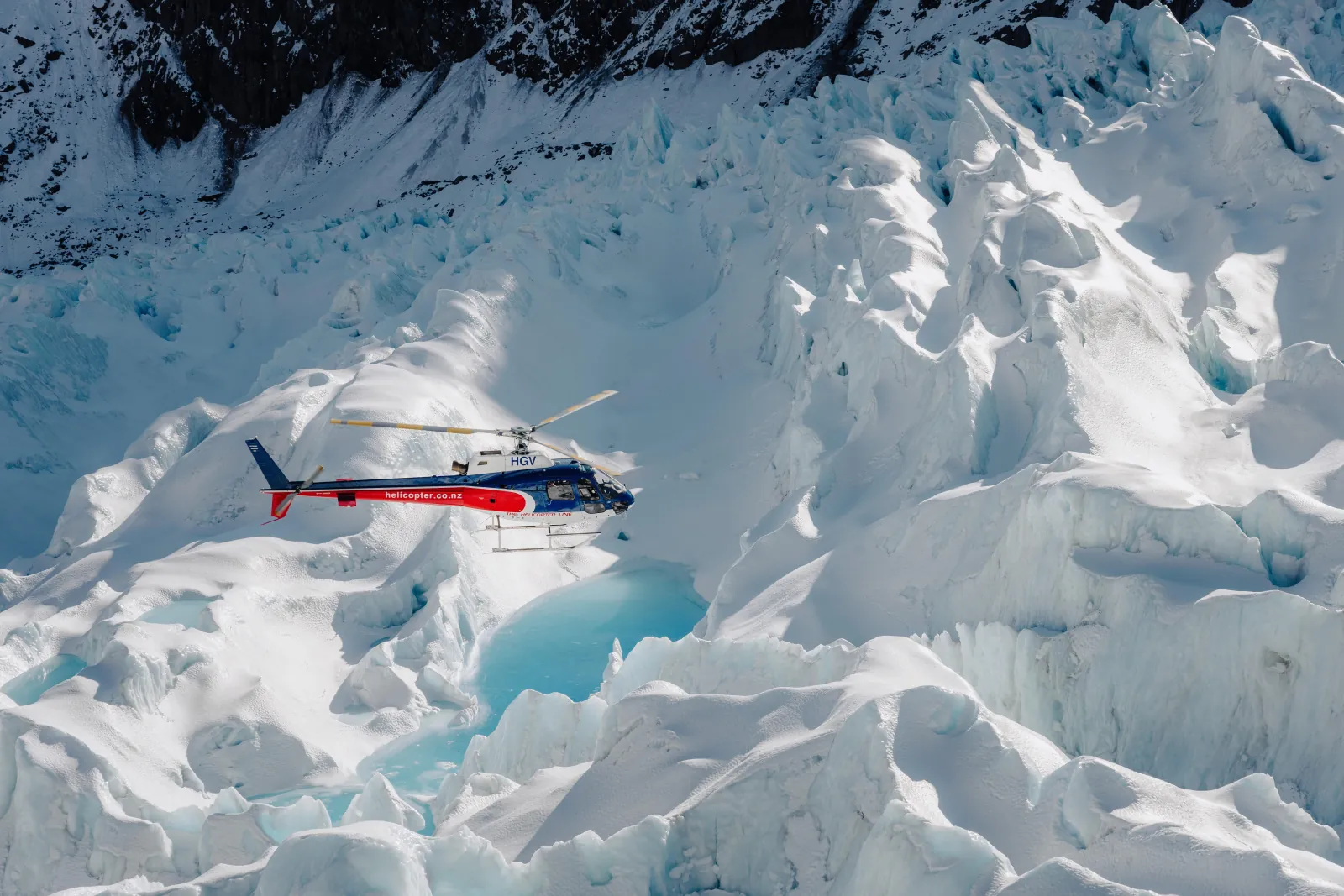 Helicopter flying over ice pinnacles and a bright turquoise melt pool on a glacier