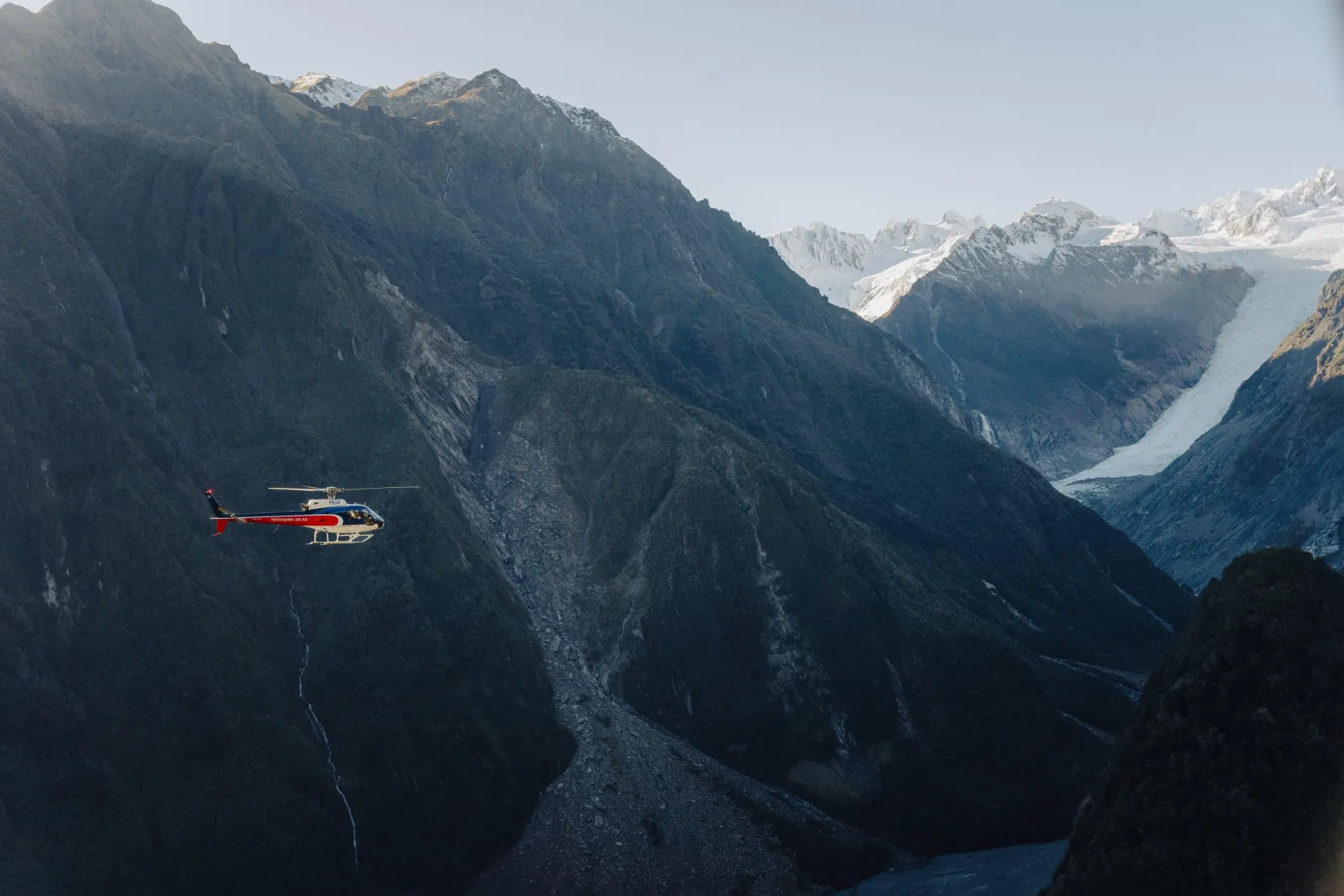 Helicopter flying low across a shadowed mountain valley with a glacier in the distance