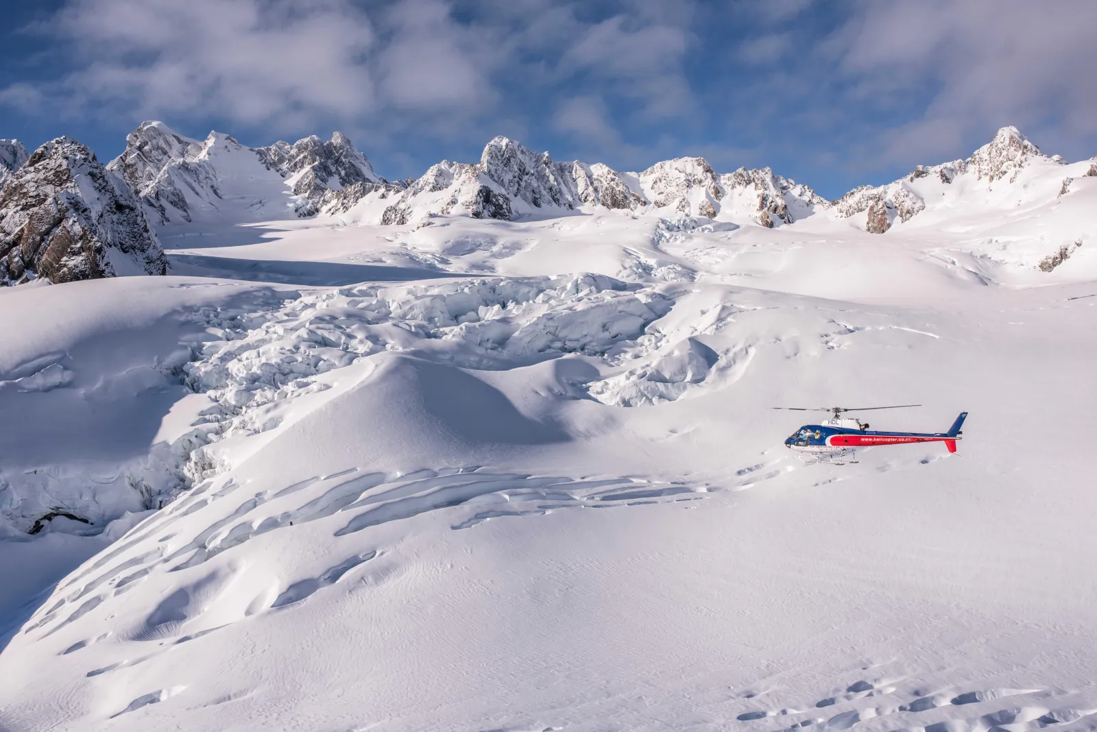 Red and blue helicopter flying low over a snow basin below jagged white peaks