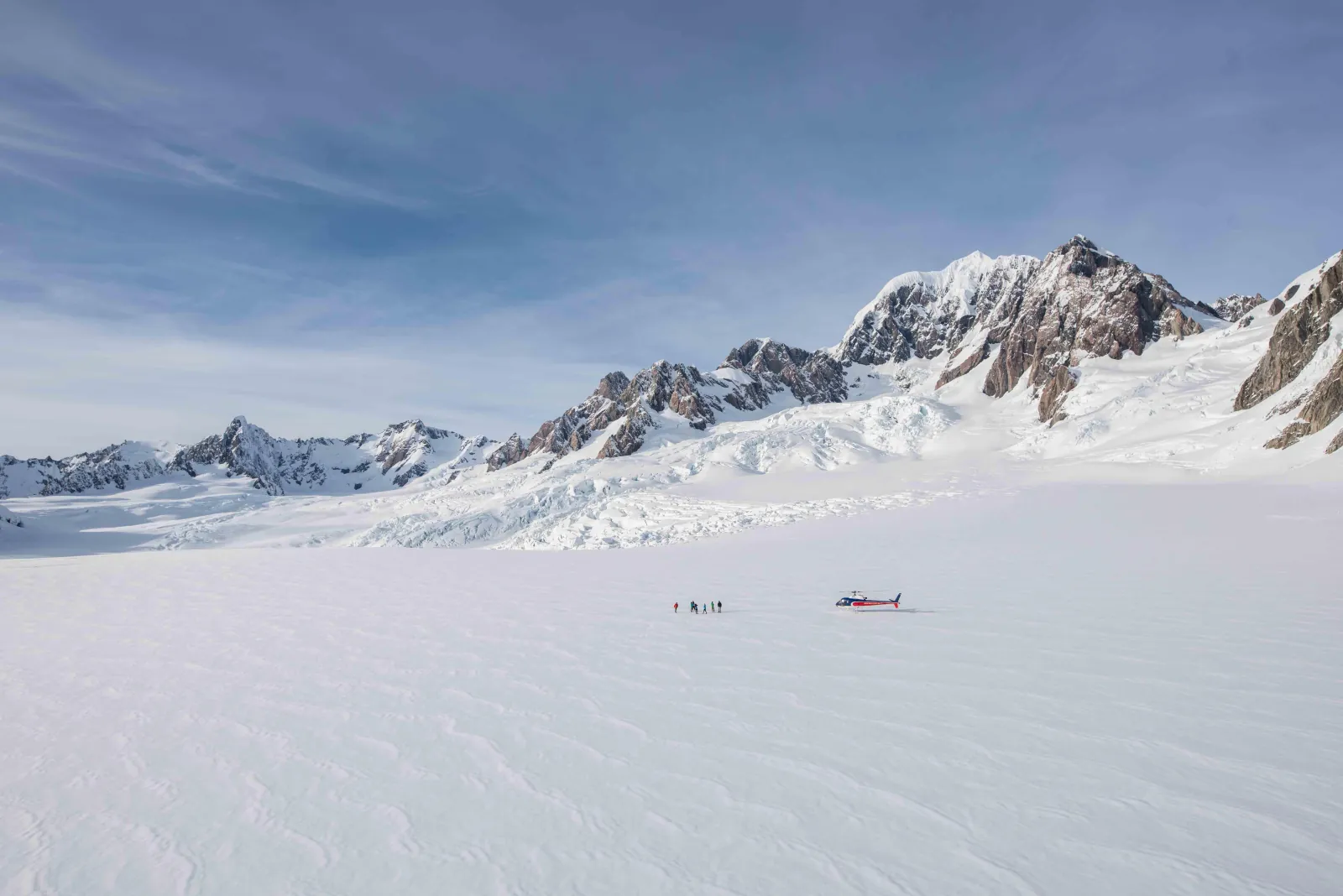 Wide snow plateau with a pair of tiny figures and helicopter before a distant ridge