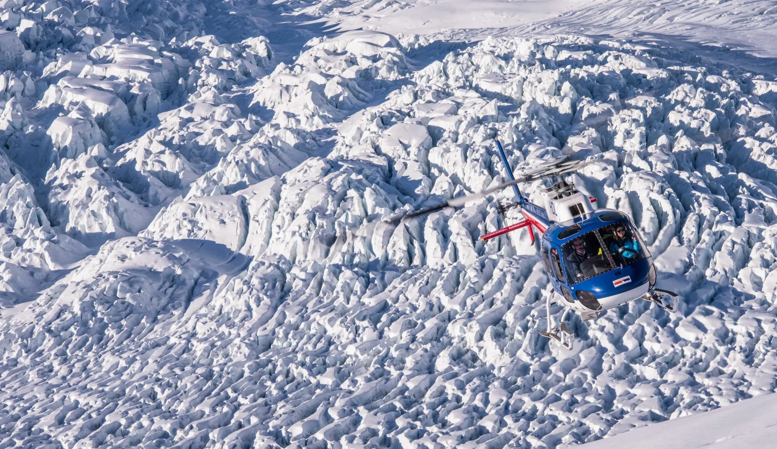 Blue helicopter hovering above a crinkled glacier surface seen from higher up