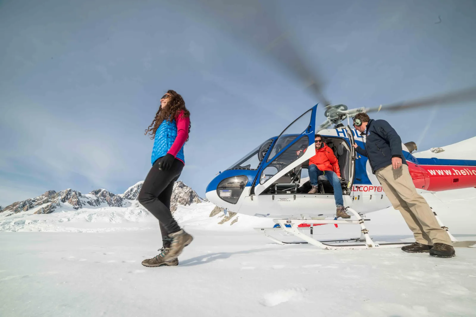 Woman striding across snow in front of a blue helicopter with passengers in its doorway
