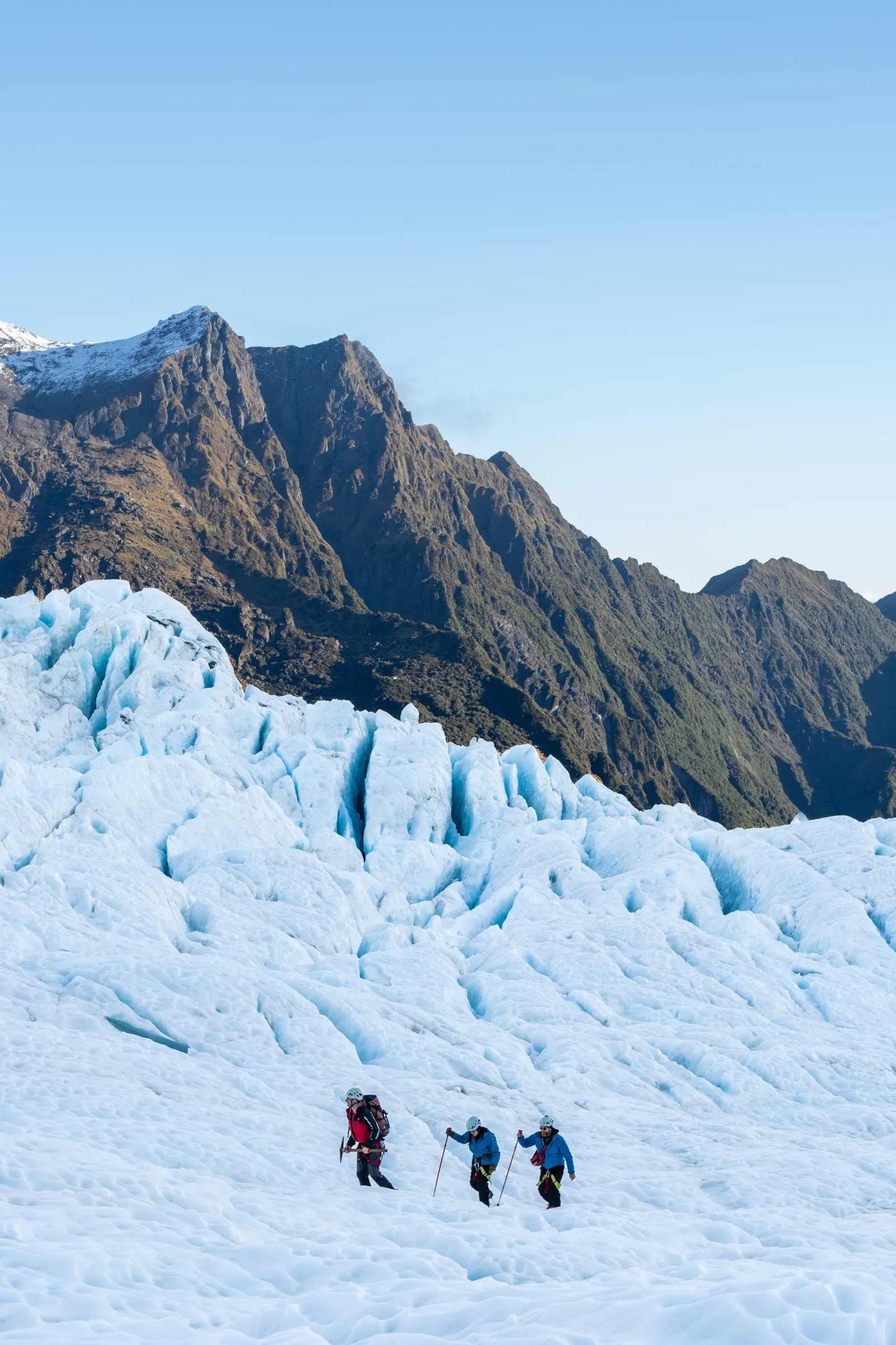Three distant hikers crossing a glacier below a mountain on a clear day