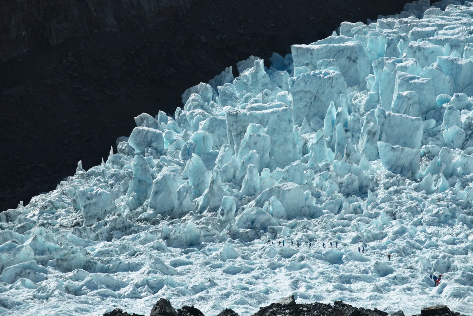 Steep dark slope covered in sharp blue-white ice towers with tiny figures far below