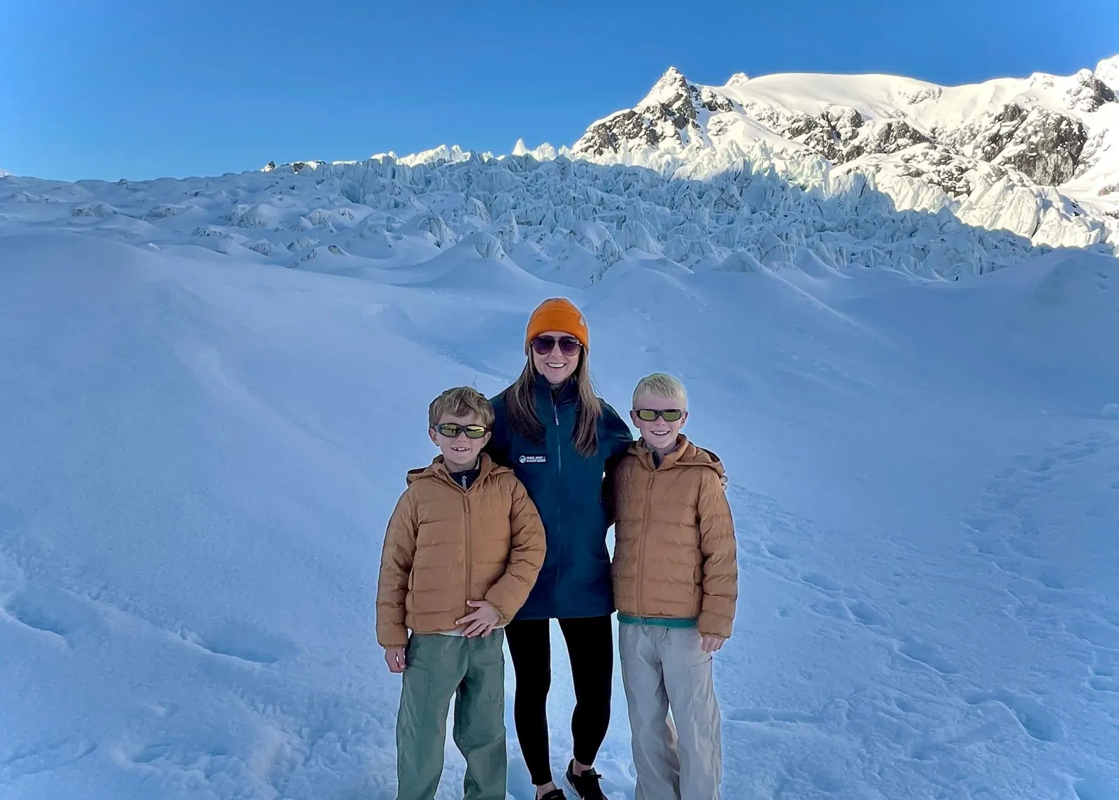 Woman in orange beanie and two boys standing on a snowfield below a peak